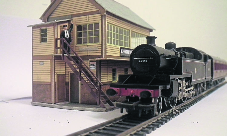 Black model train on tracks beside a model station with steps up the side and a model person stood at the top of the steps.