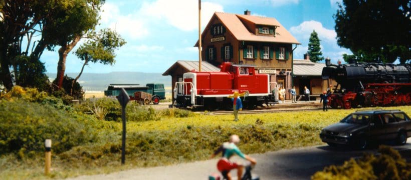 Model cyclist on path, passing in front of red model train in model railway station